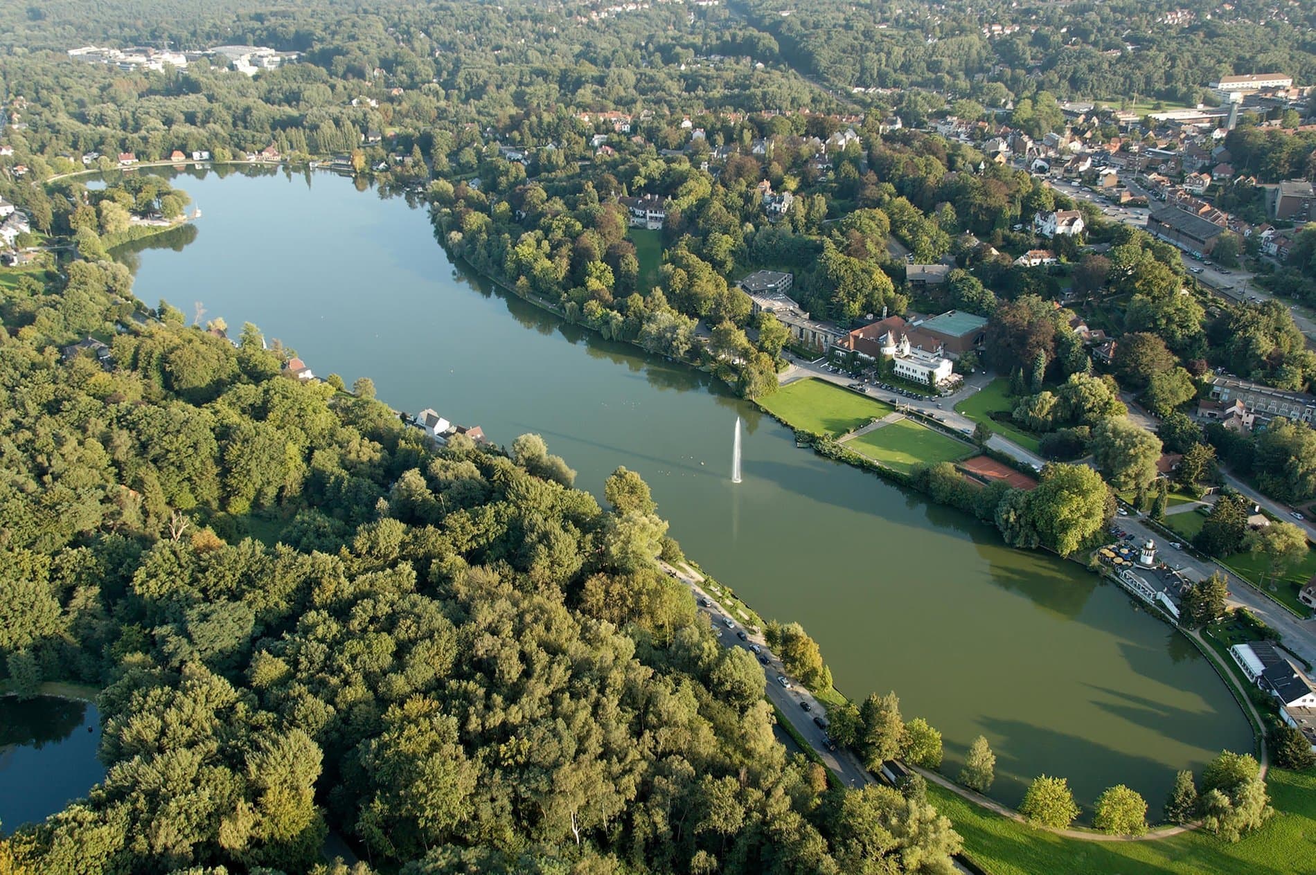 Aerial view of riverside neighborhood with dense forests, mansions, and fountain-dotted lake.