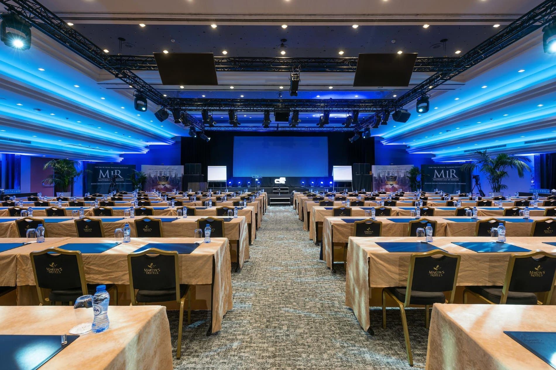 Large conference ballroom with blue ambient lighting, multiple tables, professional setup.