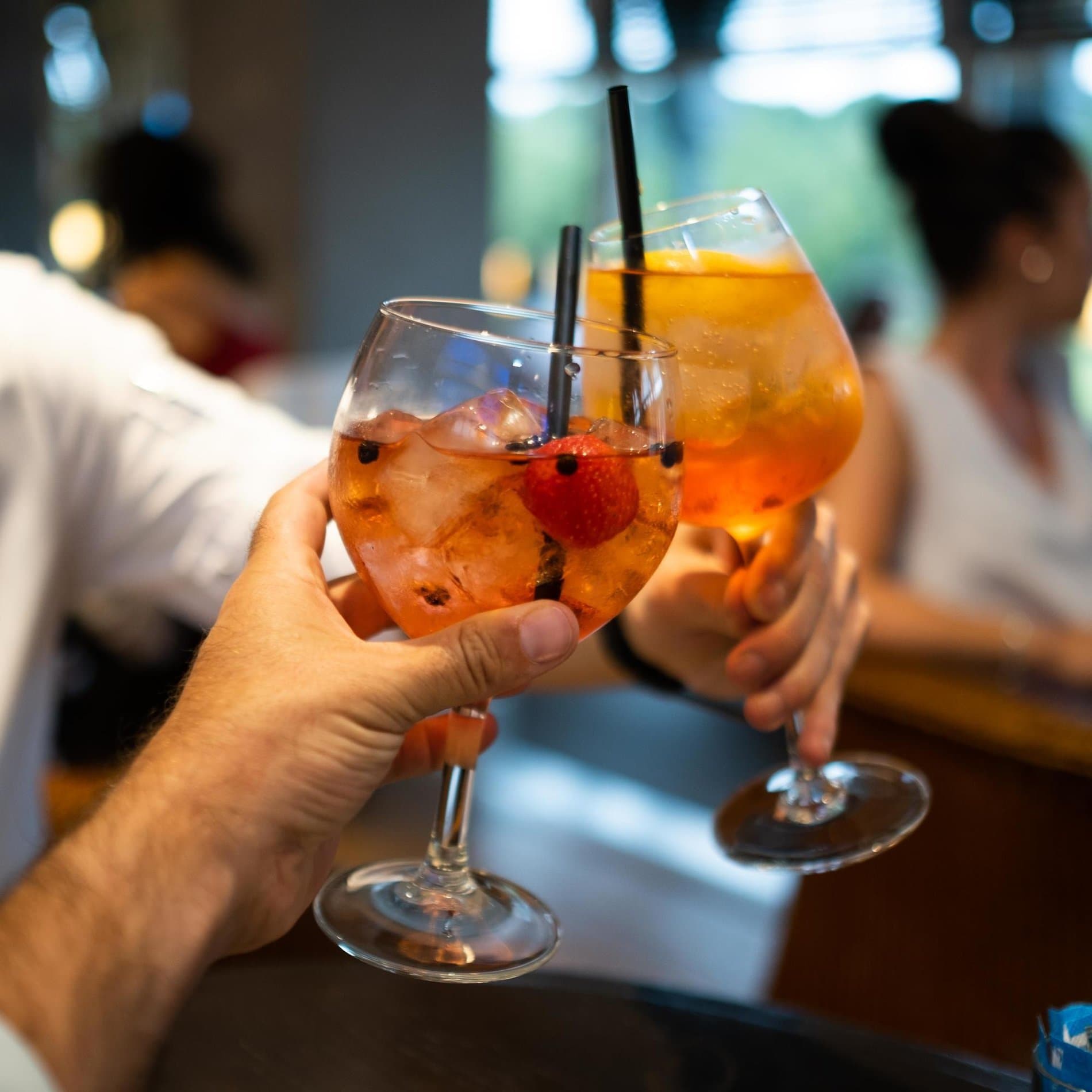 Two colorful cocktails with fruit garnish being toasted at bar area.