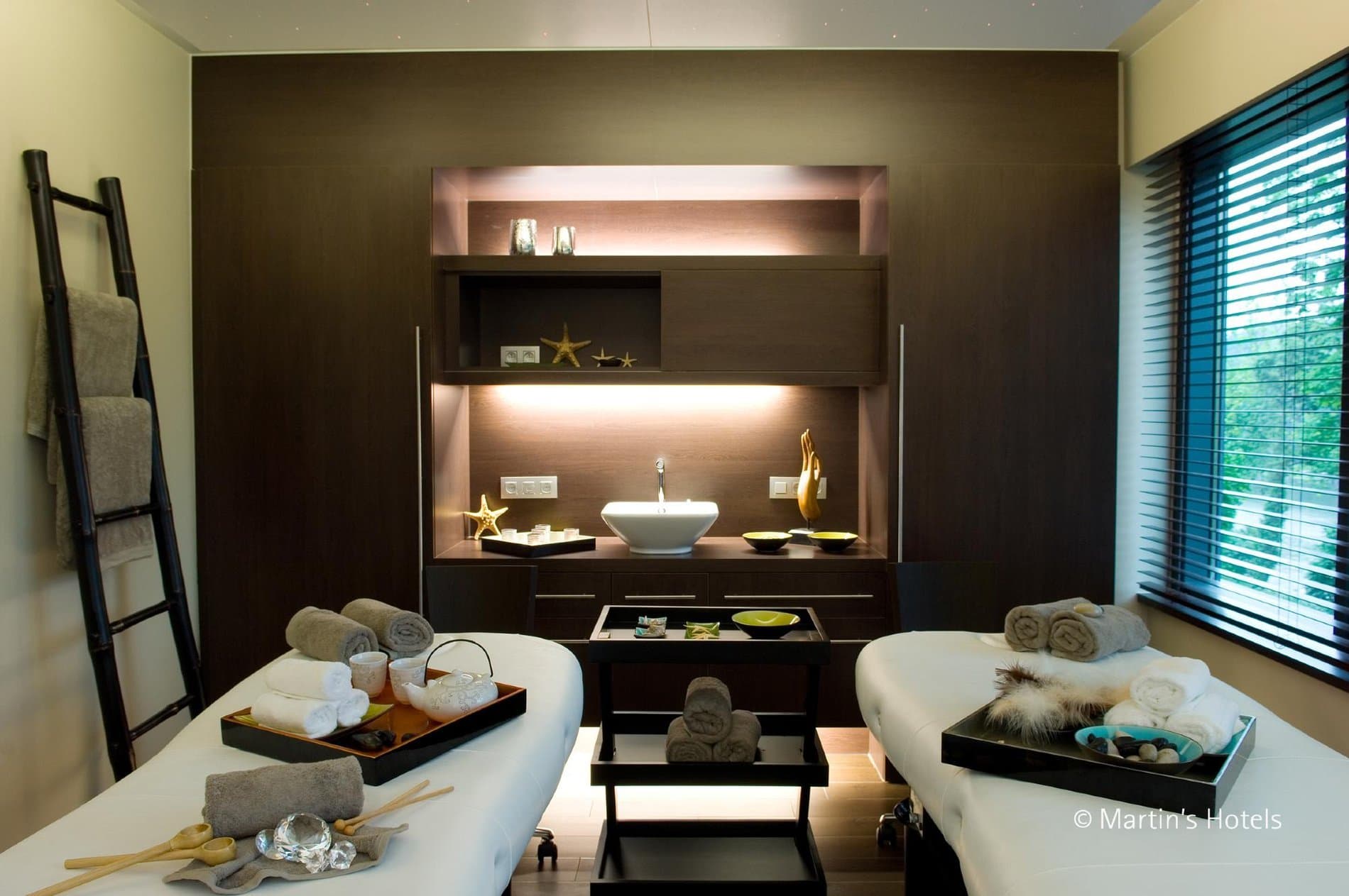 Luxury spa treatment room with dual massage beds, illuminated shelving, and decorative accents.