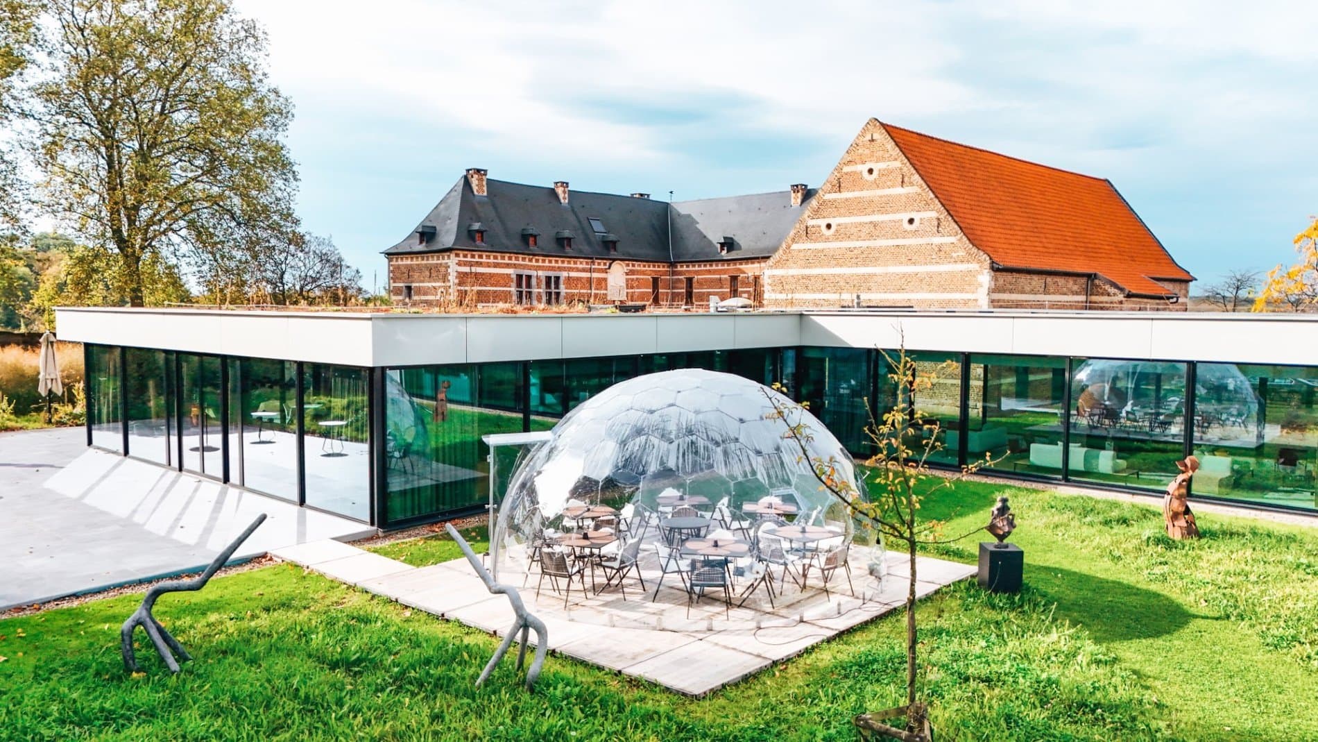 Modern glass pavilion with dome beside historic brick farmhouse and manicured gardens.