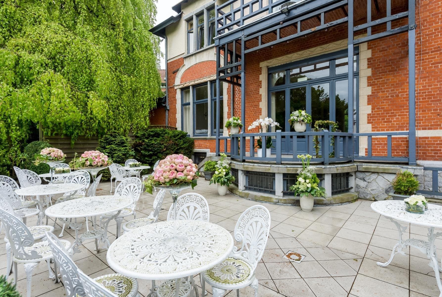 Elegant brick mansion terrace with ornate white furniture and pink flowering plants.