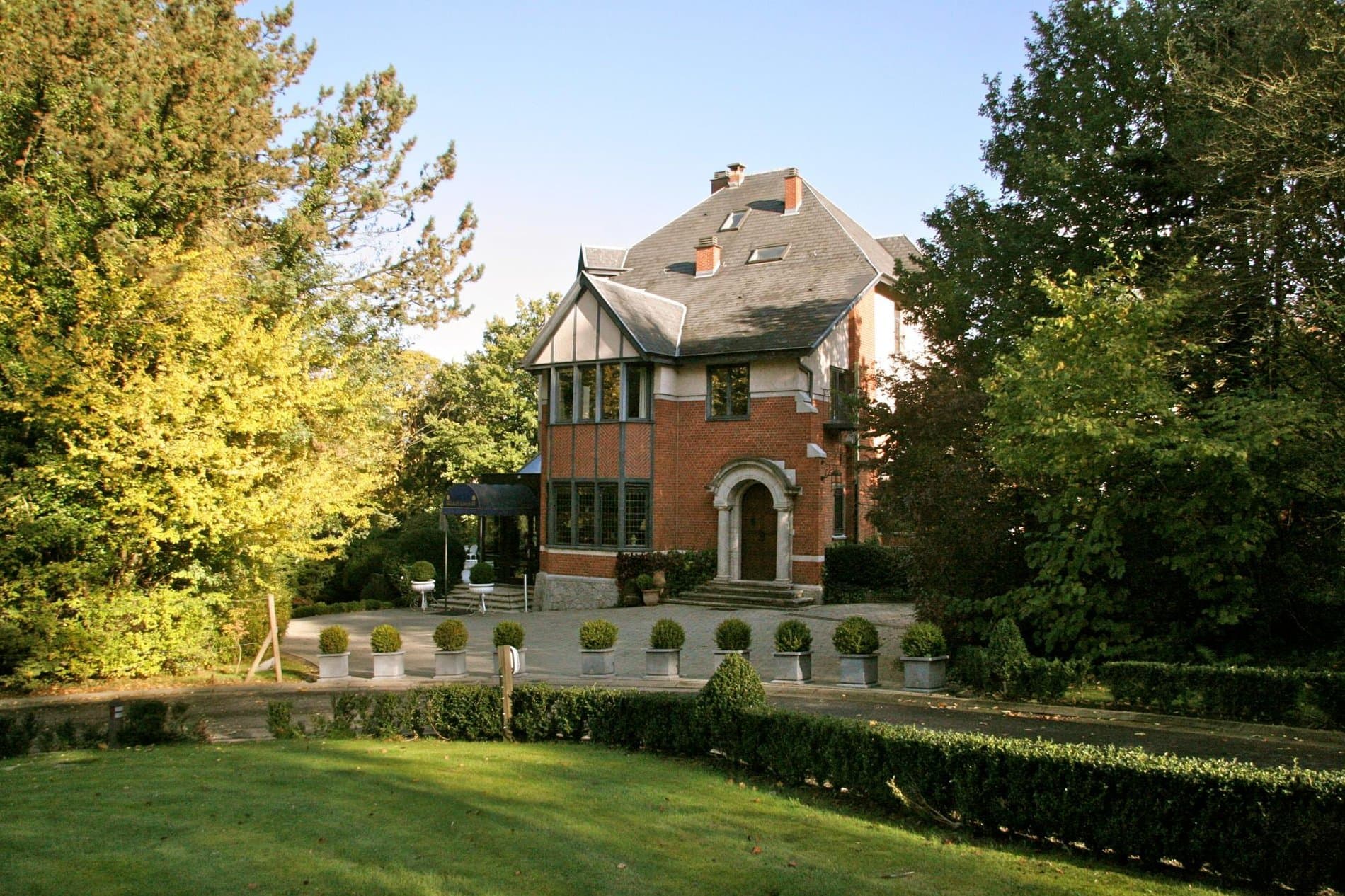 Historic brick mansion with pitched roof surrounded by manicured gardens and landscaping.