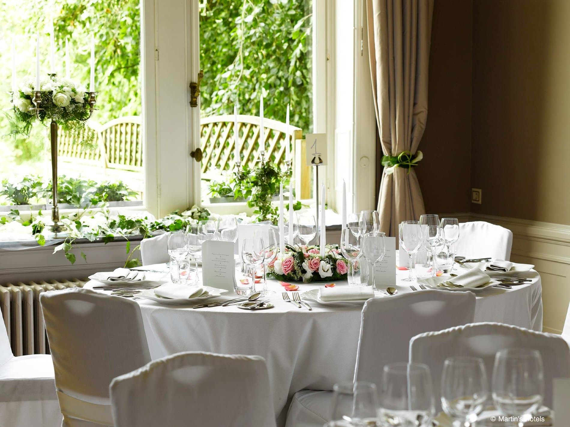 Elegant private dining room with round table overlooking lush green garden and windows.