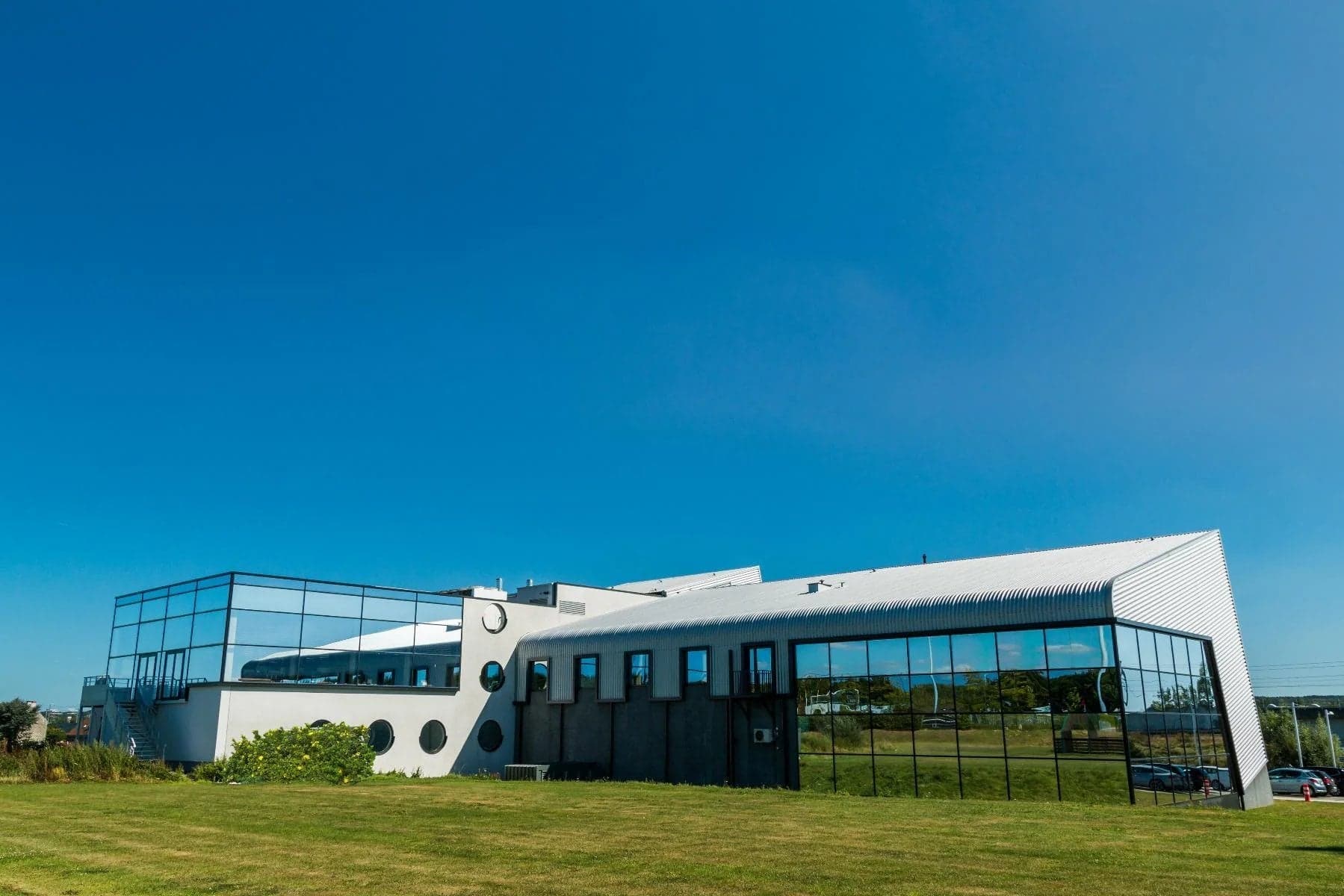 Modern curved white building with glass panels and ample parking under clear blue sky.