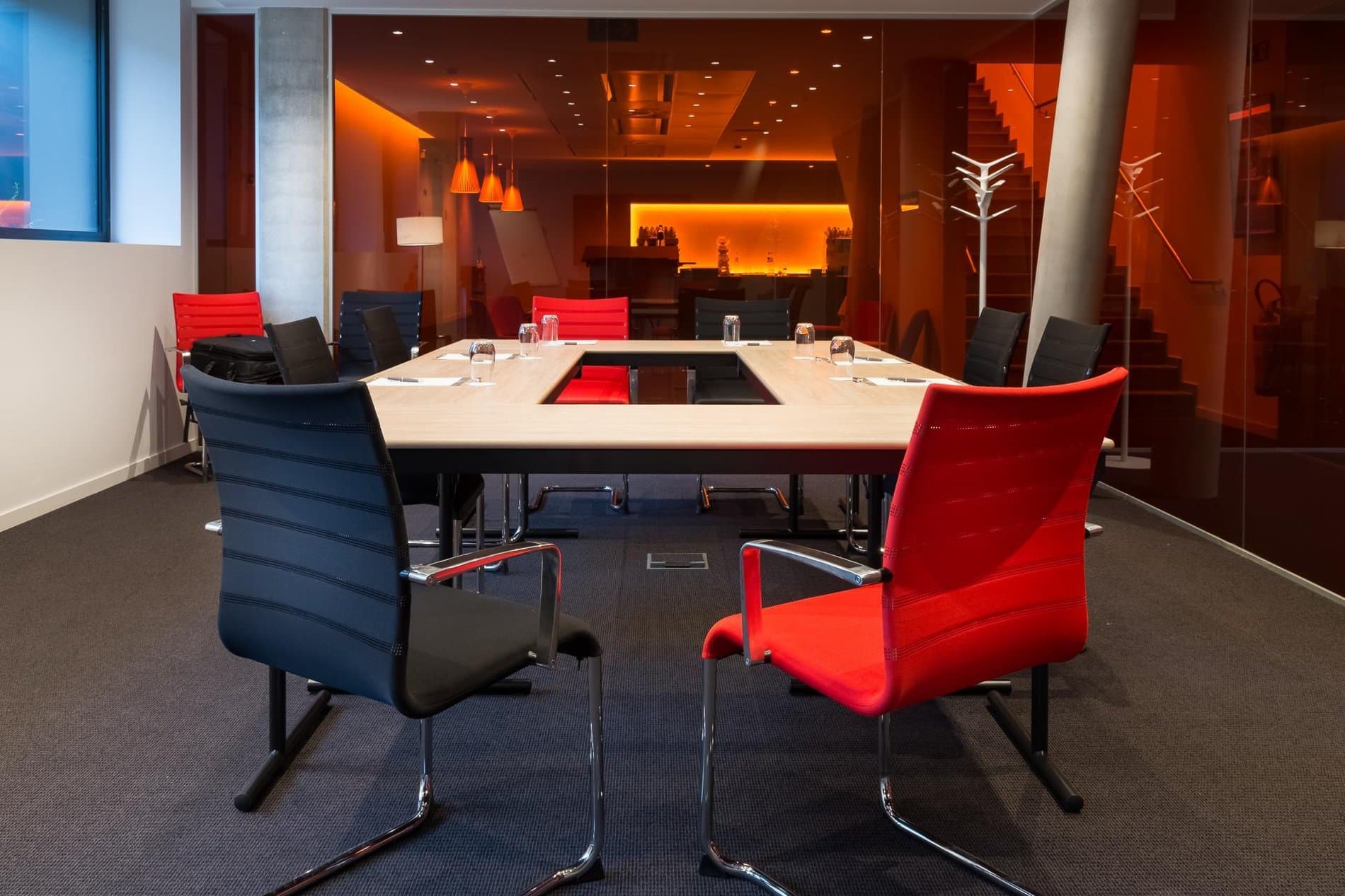 Modern conference room with red and blue chairs, orange glass walls, and minimalist furnishings.