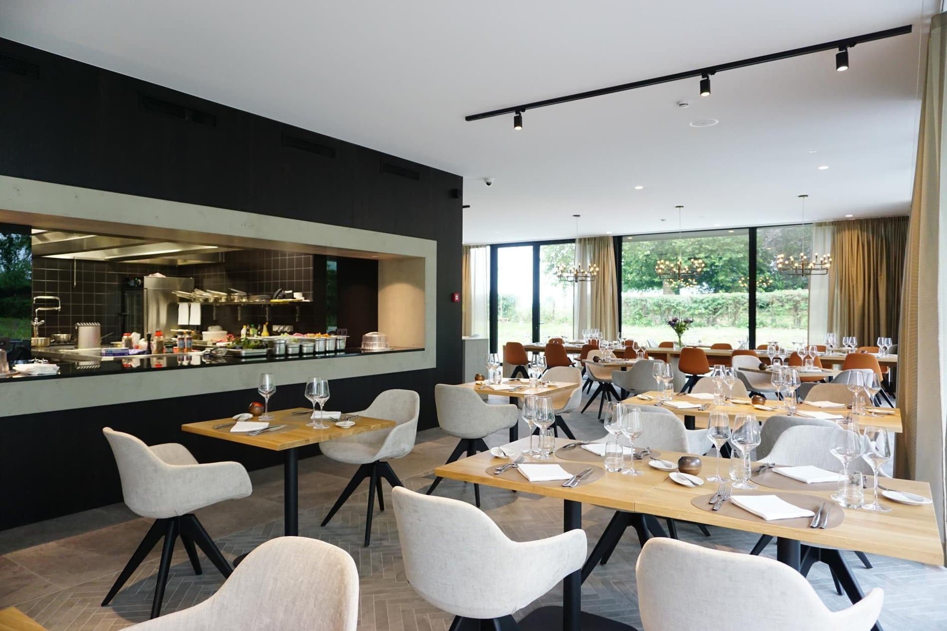 Modern restaurant with open kitchen, wooden tables, cream chairs, large windows overlooking garden.