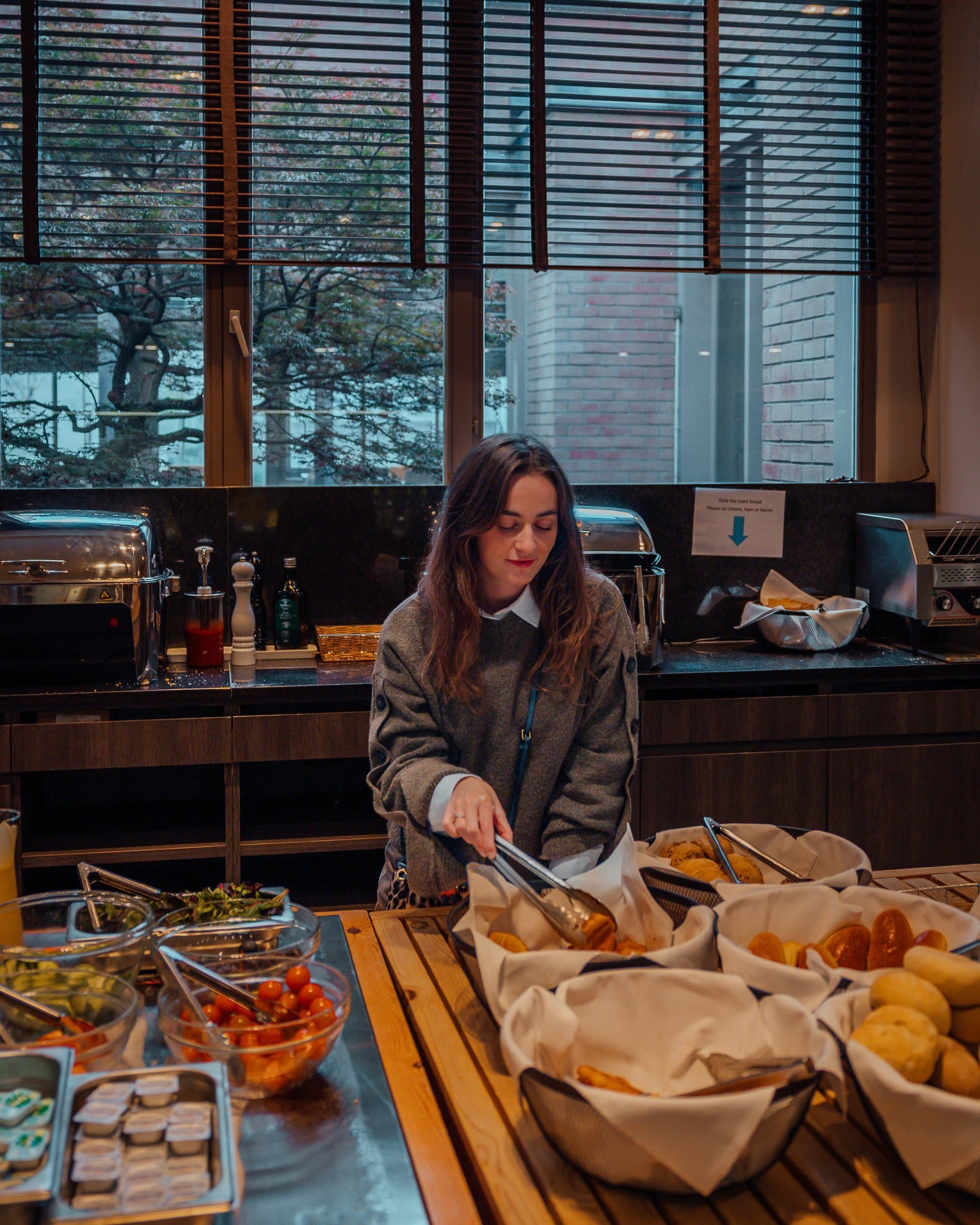 Woman serving breakfast buffet with pastries, breads, and fresh food items.