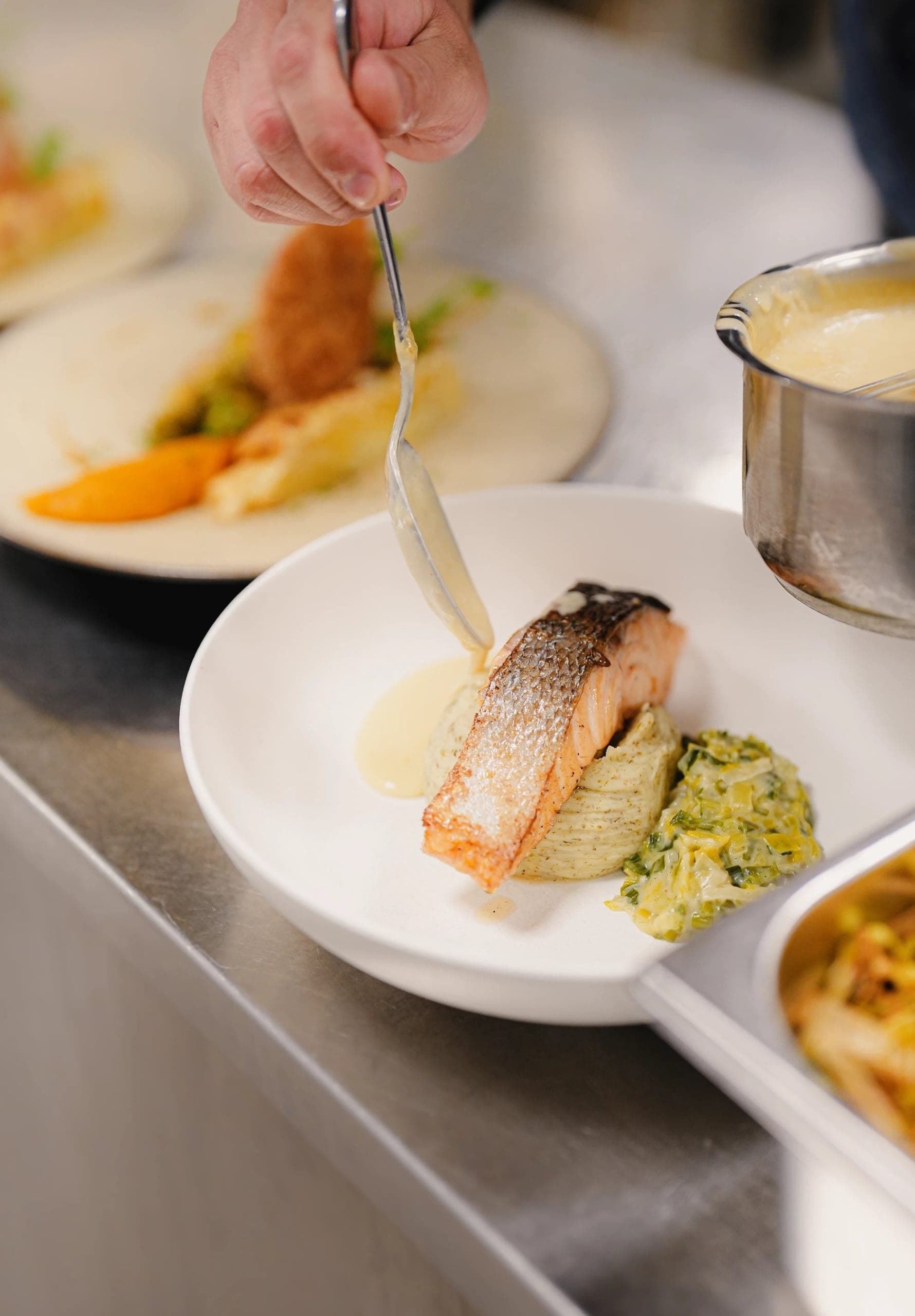 Chef plating seared salmon with sauce, vegetables, and accompaniments for diners.