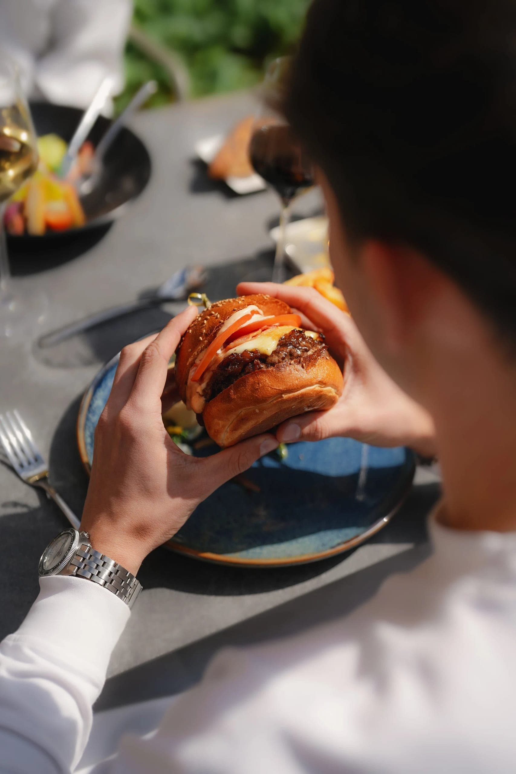 Person holding gourmet burger with wine during outdoor garden dining experience.