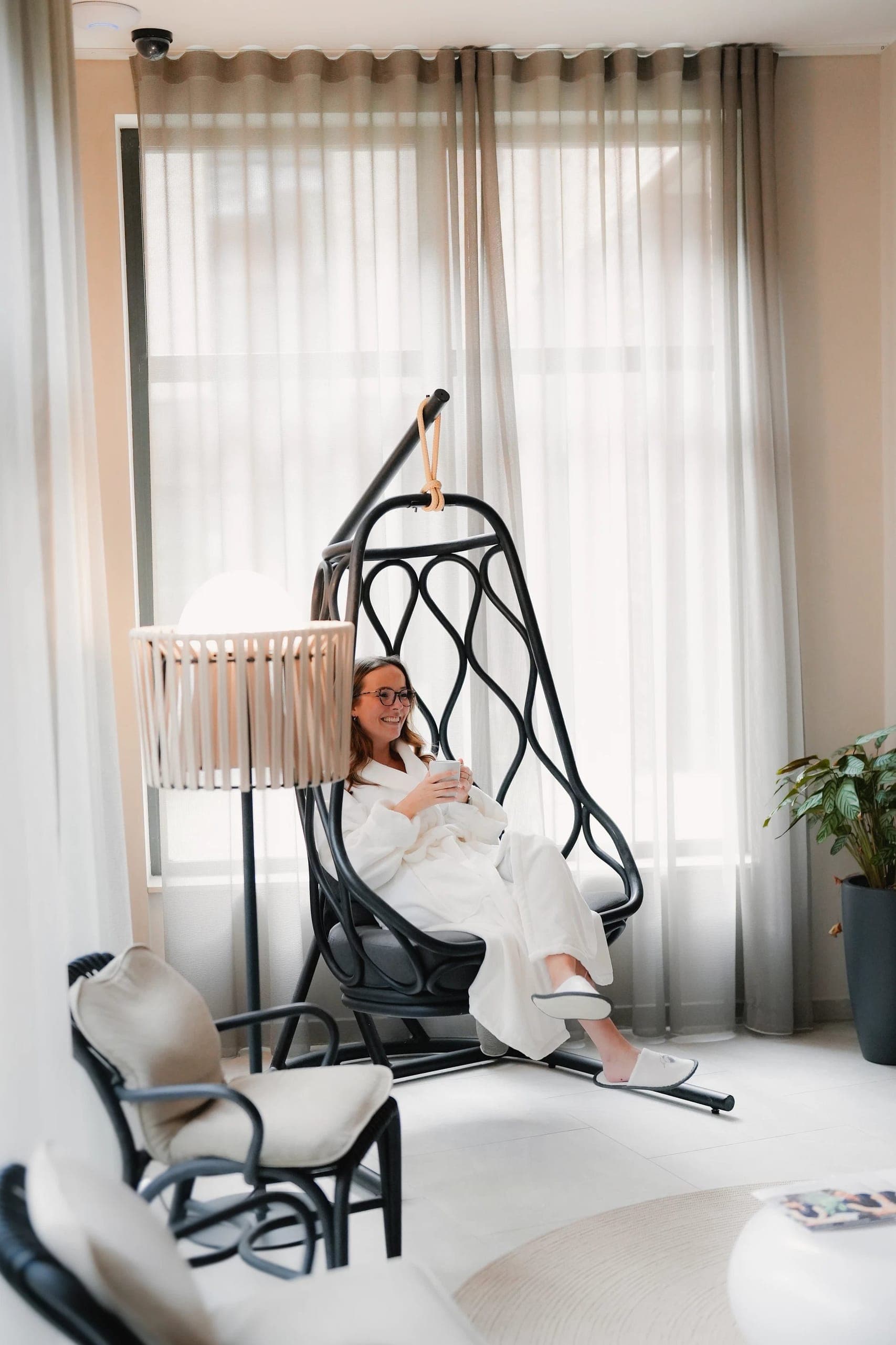 Woman relaxing in hanging chair within bright bedroom with crib and natural light.