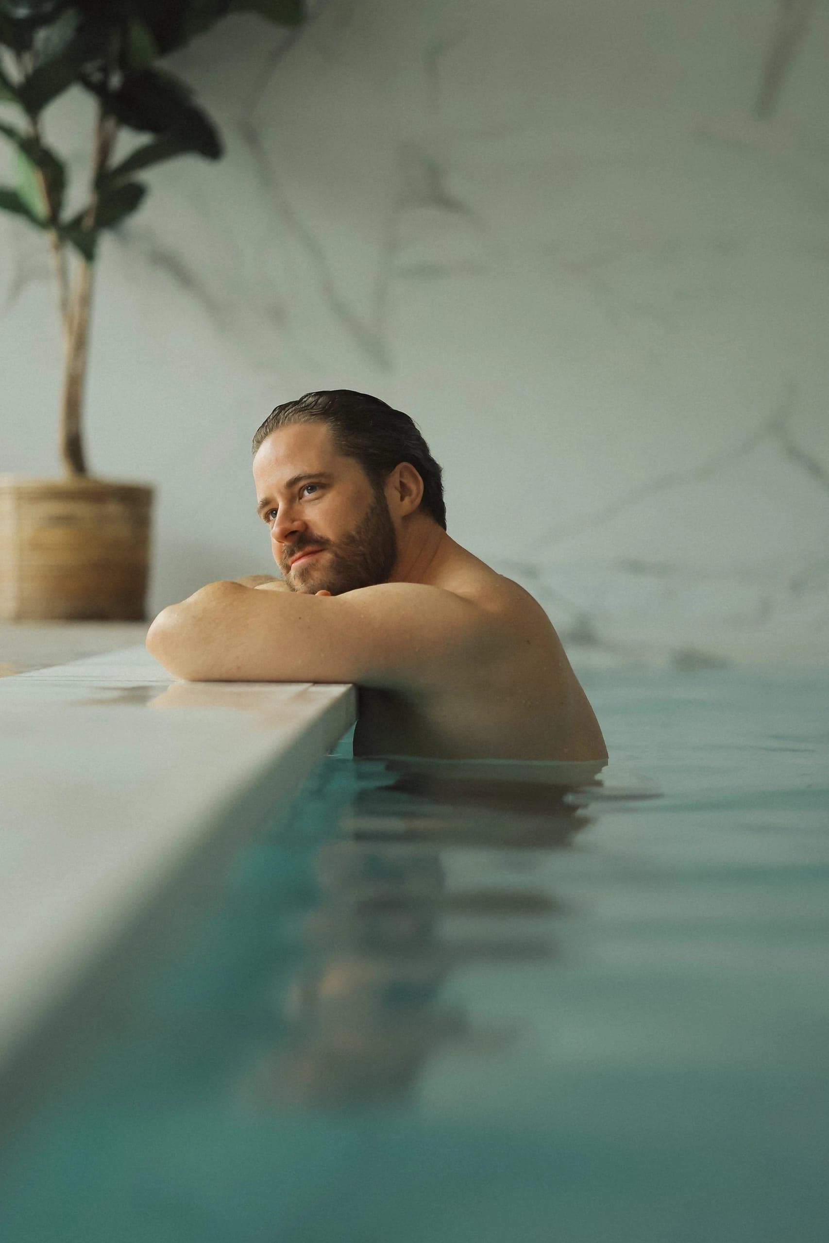 Man relaxing in indoor thermal pool with minimalist spa ambiance and potted plant.