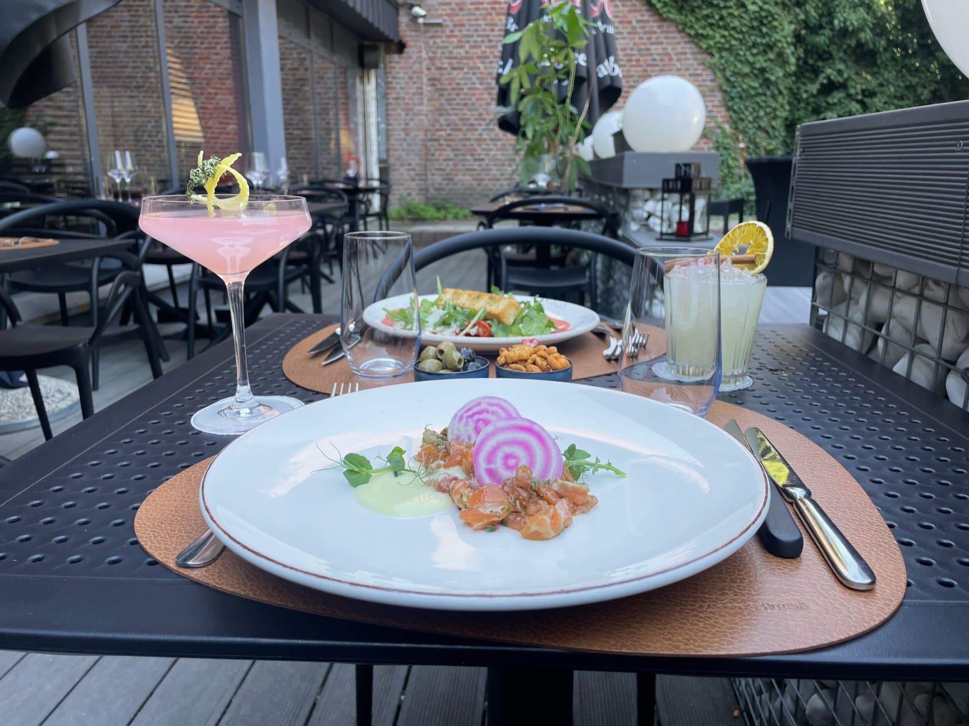 Outdoor dining terrace with plated seafood, cocktails, and ivy-covered brick building backdrop.