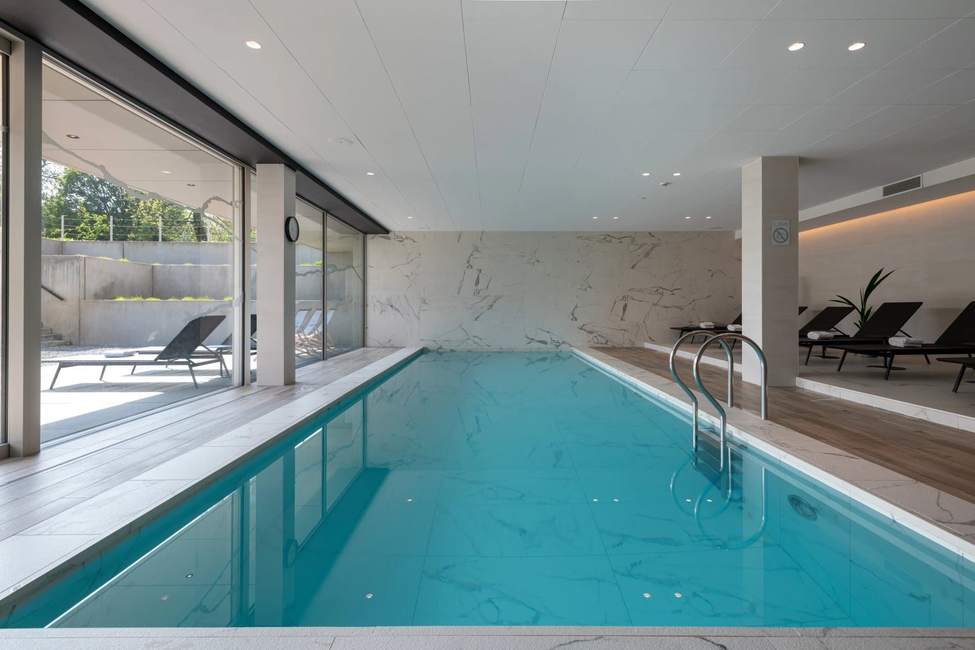 Modern indoor swimming pool with marble accents, lounge seating, and terrace access.
